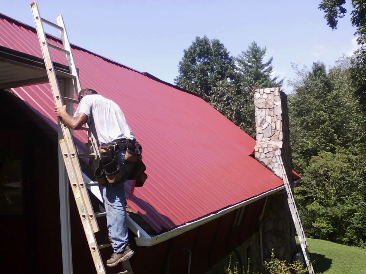 Roofer on a ladder beside a red metal roof and stone chimney — Soffit & Fascia Repair by Roofing Brothers in Almira
